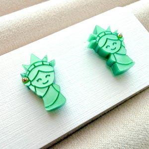 May include: A pair of light green stud earrings shaped like the Statue of Liberty, featuring a smiling face, a crown with seven points, and a small gold-toned accent on the side of the head, attached to a white rectangular display card.