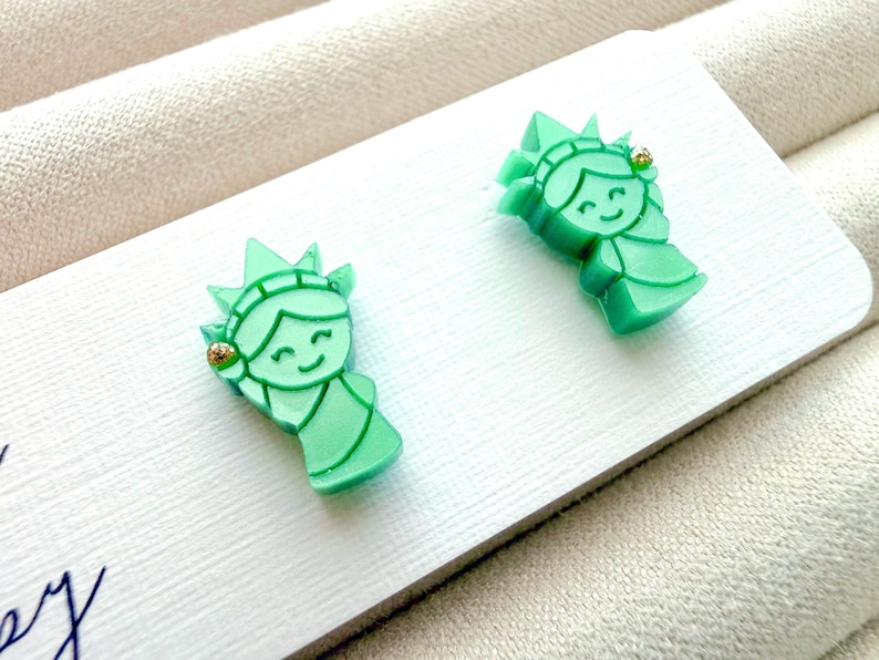May include: A pair of light green stud earrings shaped like the Statue of Liberty, featuring a smiling face, a crown with seven points, and a small gold-toned accent on the side of the head, attached to a white rectangular display card.