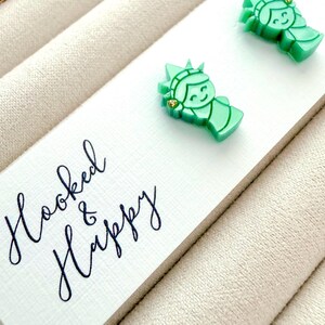 May include: A pair of light green stud earrings shaped like the Statue of Liberty, featuring a small gold-tone accent on the crown. The earrings are attached to a white display card with the black cursive text Hooked & Happy printed on it.