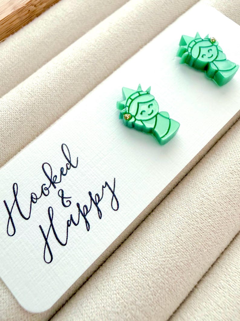 May include: A pair of light green stud earrings shaped like the Statue of Liberty, featuring a small gold-tone accent on the crown. The earrings are attached to a white display card with the black cursive text Hooked & Happy printed on it.