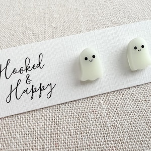 May include: A pair of white glow-in-the-dark ghost earrings with black eyes and a smile. The earrings are on a white card with the text "Hooked & Happy".