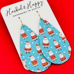 May include: A pair of teardrop-shaped dangle earrings with a blue background and a white polka dot pattern. The earrings feature a repeating pattern of Santa Claus figures in red and white.