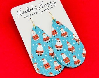 May include: A pair of teardrop-shaped dangle earrings with a blue background and a white polka dot pattern. The earrings feature a repeating pattern of Santa Claus figures in red and white.
