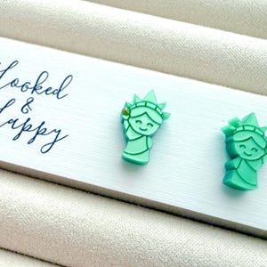 May include: A pair of mint green stud earrings shaped like the Statue of Liberty, attached to a white display card with the text Hooked & Happy printed in black script. Each earring features a small gold-tone accent on the crown.