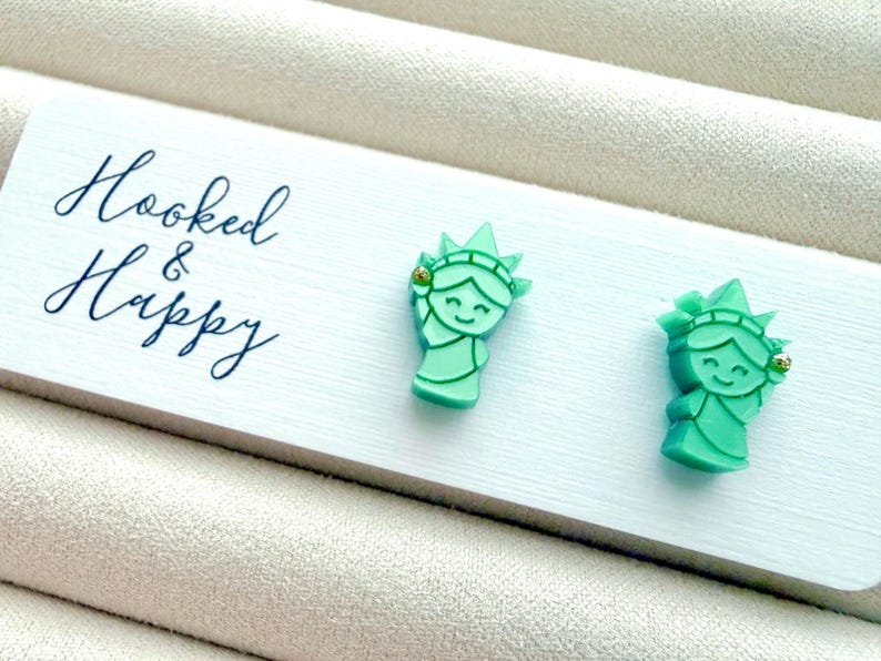 May include: A pair of mint green stud earrings shaped like the Statue of Liberty, attached to a white display card with the text Hooked & Happy printed in black script. Each earring features a small gold-tone accent on the crown.