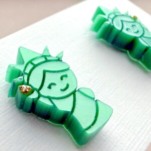 May include: A pair of stud earrings shaped like the Statue of Liberty, crafted from translucent mint green acrylic. Each earring features a small, gold-toned glitter accent on the torch, depicting the iconic figure with a smiling facial expression.