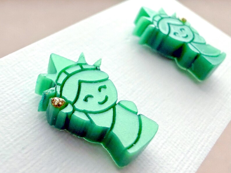 May include: A pair of stud earrings shaped like the Statue of Liberty, crafted from translucent mint green acrylic. Each earring features a small, gold-toned glitter accent on the torch, depicting the iconic figure with a smiling facial expression.