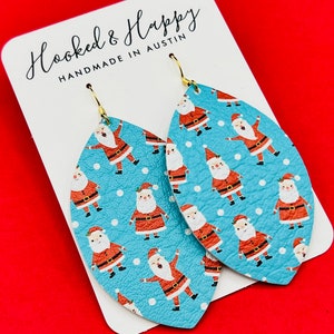 May include: A pair of blue leather earrings with a pattern of Santa Claus figures on a white background. The earrings are shaped like teardrops and have gold hooks. The earrings are handmade in Austin, Texas.