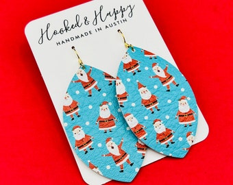 May include: A pair of blue leather earrings with a pattern of Santa Claus figures on a white background. The earrings are shaped like teardrops and have gold hooks. The earrings are handmade in Austin, Texas.