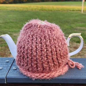 Puede incluir: Un calienta-tetera de punto de crochet rosa con una tetera blanca debajo. El calienta-tetera está hecho con un hilo esponjoso y tiene un diseño texturizado.