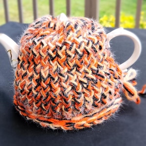 Peut inclure: Une théière blanche avec un couvre-théière en tricot orange, noir et blanc. Le couvre-théière est fait avec un fil épais et a une texture duveteuse.