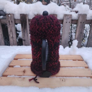 Puede incluir: Una funda de punto burdeos y negro, que cubre un objeto negro con un asa. La funda está hecha de hilo grueso y tiene un aspecto texturizado. El fondo presenta una valla de madera y nieve.