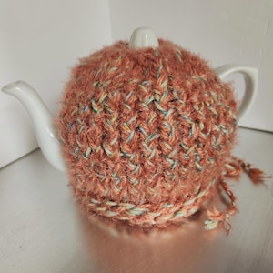 May include: A white ceramic teapot with a peach-colored knitted tea cozy. The cozy is made of a textured yarn with flecks of blue, green, and yellow. A braided trim and tassels adorn the bottom of the cozy. Tea accessories.