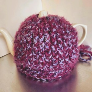 Puede incluir: Una tetera de cerámica blanca con un cubrete de punto. El cubrete es una mezcla de hilo burdeos y lavanda, que cubre la tetera. La tetera tiene un asa y un pico. El cubrete tiene un pequeño bucle de hilo adjunto.