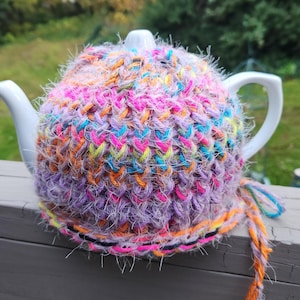 Puede incluir: Una tetera de cerámica blanca con una funda de punto hecha a mano y colorida. La funda está hecha de hilo en tonos morado, rosa, naranja, amarillo y azul. La funda tiene un aspecto texturizado y está diseñada para mantener el té caliente.