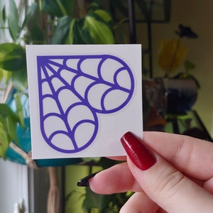 Peut inclure: Un autocollant carré avec un motif de toile d'araignée violet en forme de cœur. L'autocollant a un fond blanc et est tenu par une main avec du vernis à ongles rouge. Thème Halloween ou gothique.