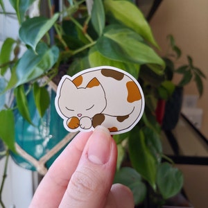 May include: A sticker featuring a cartoon cat in a sleeping pose. The cat is white with brown and orange spots, and its eyes are closed. The sticker has a black outline.
