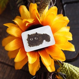 May include: A white sticker with a black cat illustration, set against a vibrant yellow sunflower. The sunflower's dark centre contrasts with its bright petals. The background is a blurred mix of dark colours and green foliage.