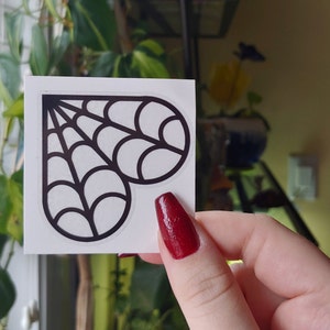 Peut inclure: Un autocollant blanc avec un motif en forme de cœur noir en forme de toile d'araignée. L'autocollant est tenu devant une fenêtre avec des plantes vertes et des stores. La personne tenant l'autocollant a du vernis à ongles rouge.