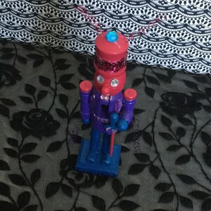 Puede incluir: Un cascanueces de madera azul y rojo con un sombrero azul y una cara roja. El cascanueces sostiene un palo rojo en su mano derecha.