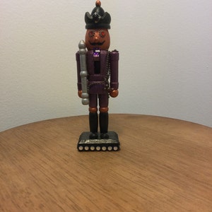 May include: A wooden nutcracker figurine with a purple and black outfit, a black hat with gold accents, and red eyes. The nutcracker is holding a silver-colored staff and is standing on a black base with silver accents.