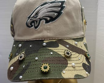 Może przedstawiać: Beżowa i moro czapka bejsbolowa z logo Philadelphia Eagles. Czapka jest ozdobiona dżetami, perłami i dekoracyjnymi guzikami z zielonymi i czarnymi akcentami. Daszek jest moro.