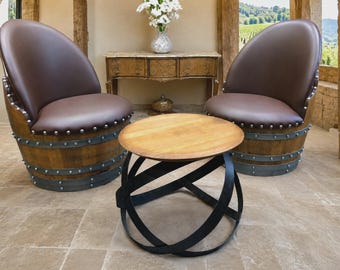 May include: Two armchairs constructed from wooden barrels with dark brown upholstered seats and silver nailhead trim, paired with a round wooden coffee table featuring a black metal base made of intersecting curved bands.
