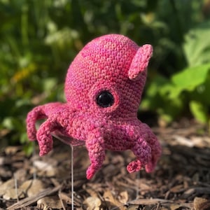 May include: A handmade, pink crocheted octopus with a dark eye and small ear-like appendages. The octopus has eight tentacles and is displayed on a clear stand against a blurred green and brown background. The yarn has a speckled appearance.
