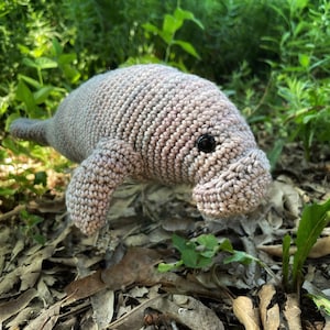 May include: A crocheted manatee plush toy with a pink and gray color scheme. The toy has a black button eye and is posed on a bed of brown leaves.
