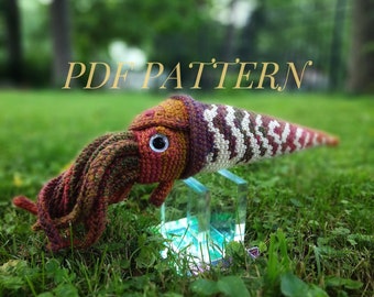 May include: A crocheted amigurumi squid with a long, striped body and a colorful, textured head. The squid is sitting on a clear glass stand. The text "PDF PATTERN" is visible in the background.