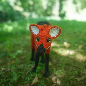 Puede incluir: Un animal de peluche de crochet que se asemeja a un zorro rojo con detalles en blanco y negro. El animal tiene ojos verdes y una nariz negra. El animal está de pie sobre un lecho de hierba verde.