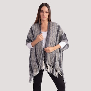 May include: A grey and beige woven poncho with fringe detailing. The poncho features a striped pattern and is worn over a white long-sleeved top and black trousers. The garment has a relaxed fit and a textured appearance.