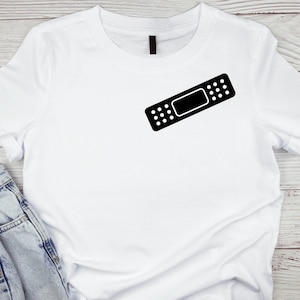 May include: White t-shirt with a black band-aid graphic on the front. The t-shirt is laying flat on a wooden surface.