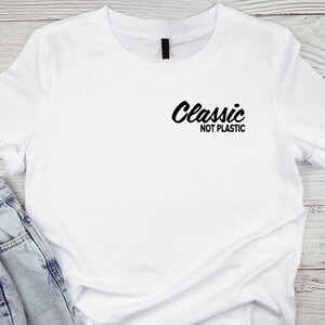 Puede incluir: Camiseta blanca con la frase "Classic NOT PLASTIC" en negro. La camiseta es de manga corta. Unos vaqueros azules son parcialmente visibles en la imagen.
