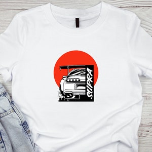 Puede incluir: Camiseta blanca con una ilustración en blanco y negro de un coche Supra delante de un círculo rojo. El texto "SUPRA" está escrito verticalmente en el lado derecho del coche.
