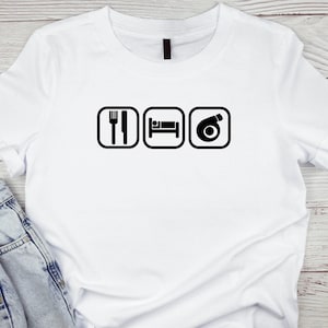 Puede incluir: Camiseta blanca con un diseño gráfico negro. El diseño presenta tres iconos cuadrados negros: un tenedor y un cuchillo, una cama y un turbocompresor. La camiseta se combina con unos vaqueros azules.