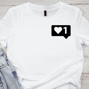 Puede incluir: Camiseta blanca con un gráfico negro de un icono de notificación de redes sociales con un corazón blanco y el número "1" dentro de un bocadillo.