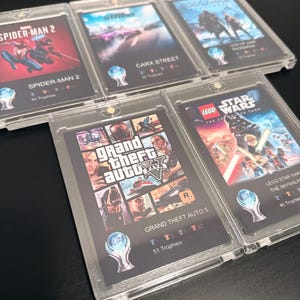 Può includere: Una collezione di custodie di videogiochi, tra cui "Spider-Man 2", "Carx Street", "God of War Ragnarok", "Grand Theft Auto 5" e "Lego Star Wars: The Skywalker Saga". Ogni custodia presenta grafica di gioco e conteggio dei trofei, protette da copertine trasparenti.