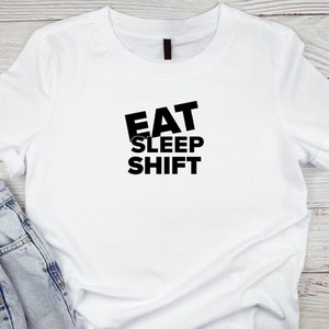 Puede incluir: Camiseta blanca con texto negro que dice "EAT SLEEP SHIFT" sobre un fondo de madera blanco.
