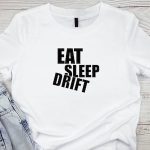 Puede incluir: Camiseta blanca con las palabras negras "EAT SLEEP DRIFT". La palabra "DRIFT" está en ángulo. La camiseta se combina con unos vaqueros azules. La camiseta está hecha de un material suave.