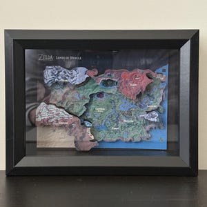 Peut inclure: Carte 3D encadrée de Zelda: Lands of Hyrule, représentant la topographie détaillée du monde du jeu. La carte est multicolore, avec différentes régions étiquetées, et est encadrée en noir.