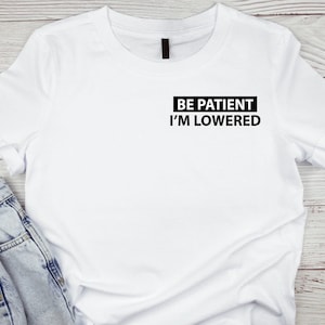 Puede incluir: Camiseta blanca con texto negro que dice "BE PATIENT I'M LOWERED" en la parte delantera.
