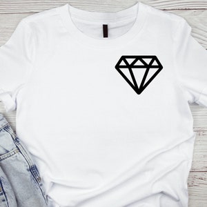 Puede incluir: Camiseta blanca con un gráfico de diamante negro en la parte delantera. La camiseta se muestra con vaqueros de mezclilla azules. El diseño del diamante es una forma geométrica simple.
