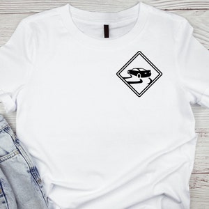 Puede incluir: Camiseta blanca con un gráfico negro de un coche conduciendo por una carretera resbaladiza, dentro de una señal de advertencia en forma de diamante.