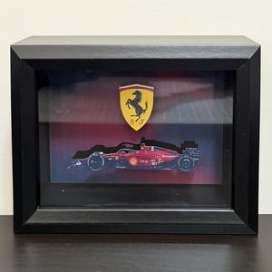Puede incluir: Obra de arte enmarcada con un coche de carreras Ferrari de Fórmula 1 y el emblema de Ferrari. El coche rojo, con logotipos de patrocinadores, se encuentra sobre un fondo degradado rojo y azul. El marco negro añade un acabado elegante.