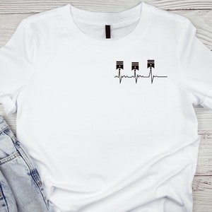 Puede incluir: Camiseta blanca con un gráfico negro de tres pistones y una línea de latido del corazón.