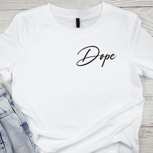 Puede incluir: Camiseta blanca con la palabra "Dope" en escritura negra en el pecho. La camiseta se combina con vaqueros azules. La camiseta tiene mangas cortas y cuello redondo.