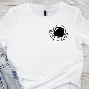 Puede incluir: Camiseta blanca con un diseño de astronauta de dibujos animados en blanco y negro. El astronauta tiene los brazos levantados. La camiseta se combina con vaqueros azules, lo que sugiere ropa informal.
