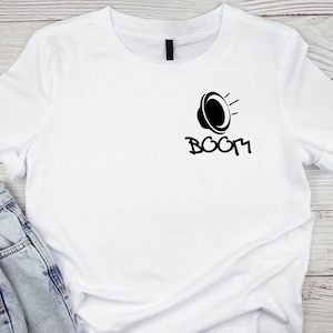 Puede incluir: Camiseta blanca con un gráfico negro de un altavoz y la palabra "BOOM" en fuente de grafiti. La imagen incluye vaqueros azules, lo que sugiere un estilo casual y cotidiano.