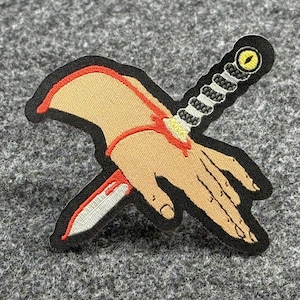 Könnte beinhalten: Ein gestickter Patch in Schwarzweiß, der eine Hand mit einem Messer zeigt, das durch sie hindurchgeht. Die Hand ist hautfarben und das Messer hat einen gelben und schwarzen Griff.